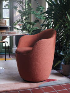 A modern muted brick-red curved armchair with green plants to the right.