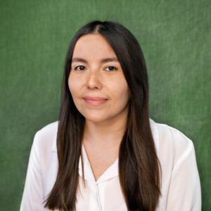 Headshot portrait of woman with long brown hair, wearing a white shirt in front of a forest green background.