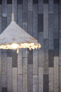 Rectangular tiles, of various grey hues, line a wall behind a blurry off-white hanging lamp.