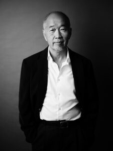 A black and white headshot image of a man looking to camera wearing a blazer and a white button-up shirt.