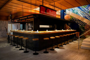 A restaurant bar is shown, with a light wood tabletop and stools, with black accents.