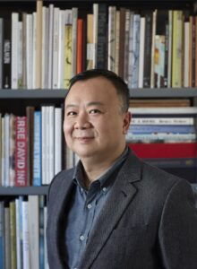 A headshot of a man in a button up shirt and suit jacket in front of a bookcase filled with books.