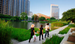People walk on a sidewalk along water amongst abundant greenery and city buildings in the background.