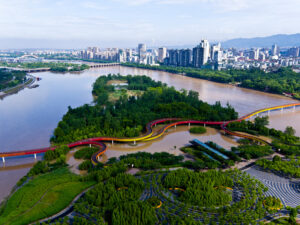 An aerial view of a triangular park surrounded by a brown river with a city skyline and mountains in the background and a red and yellow walkway running horizontally.