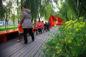 Children line up behind an adult on a paneled walkway with bright red seat and sculptural accents.