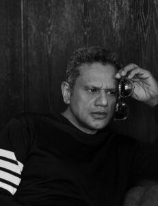 A black and white headshot image of a man wearing a black shirt with his hand to his forehead holding eyeglasses.