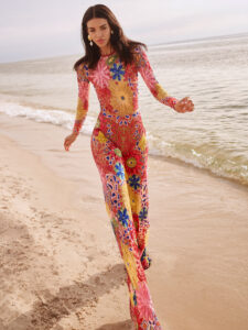 A person wears a pink, yellow, and blue bodysuit while walking on sand along a beach.