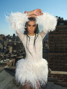 A person stands with their arms resting on their head, wearing an all-white dress with beading on the torso and thin feathers on the arms and bottom of the dress; a city skyline is in the background.
