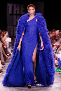 A model on a runway wears a royal blue one-shoulder floor-length dress with a feathered cape of the same color.