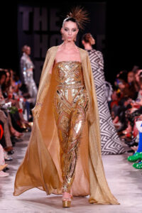 A model on a runway wears a shiny gold detailed one piece fitted outfit, with a flowing gold cape and long gold beaded gloves.