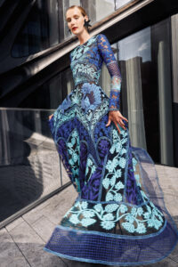 A model looks down at the camera; she wears a long-sleeve floor length dress with intricate cut-out details in black, blue, and aqua colors.
