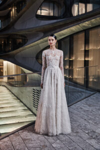 An individual wears a long sleeve floor length white dress that is semi-sheer and has intricate details; they stand in front of dramatic curved architecture.