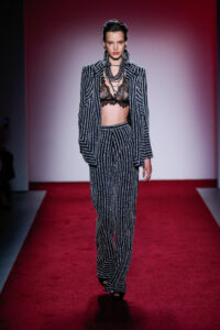 A runway model wears black trousers and a blazer, both detailed with vertical-lined silver beading.