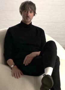 A man wearing a black turtleneck shirt and black pants sits on a plush white chair.