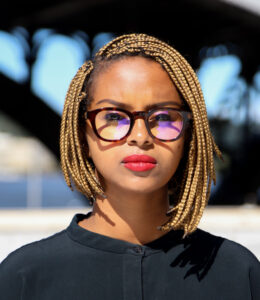 A headshot image of a woman wearing a black shirt, brown glasses, and red lipstick.