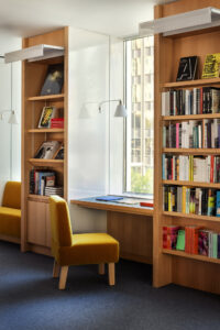 An interior featuring two vertical bookshelves, in the center is a desk with a yellow plush chair facing a window.