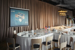 A conference room interior with long tables topped with a glass surface, surrounded by chairs; long beige curtains are the backdrop.