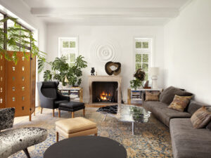 A sitting room with black and grey couches and seats, with a lit fireplace, surrounded by some green decorative plants.