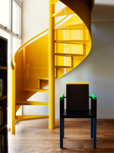 A bright yellow spiral staircase with a modern style black chair with yellow and lime green accents on the right.