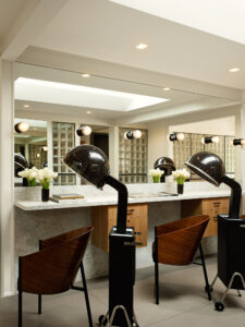A modern interior of a salon with dark wood chairs and black standing hair dryers in front of a large mirror.