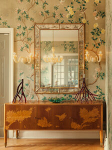 A brown detailed dresser sits below a large mirror, behind is an elaborate wallpaper with white flowers and green leaves.