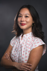 A headshot image of a woman wearing a patterned white shirt with small red details, arms folded, looking to camera.