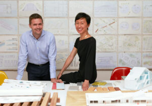 A man stands behind a large table covered with papers and items, and a woman to the right sits on the desk, both smiling to camera.