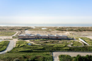 An aerial photograph of a long rectangular building surrounded by grass in the foreground and grassy and sandy beach and water in the background.