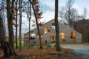 A home with a triangular roof, a light wooden exterior, and several large square windows sits within trees with leaves on the ground.