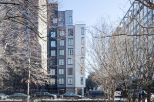 A photograph of a car-lined street with a vertically rectangular building of decreasing heights featured at center, in grey-scale hues; tree branches are in the foreground on both sides.