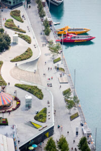 An aerial view of a waterside park, with boats on the water to the right and people walking on a sidewalk amongst grassy sections to the left.