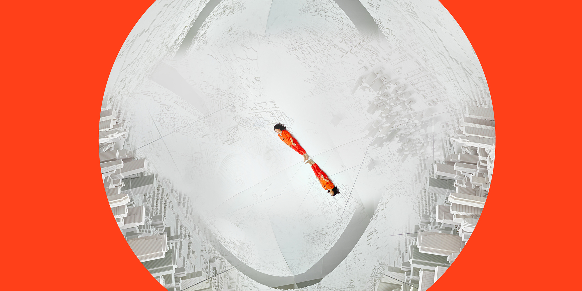 Es Devlin, a white woman with dark hair wearing a vibrant red jumpsuit, appears miniscule in the center of a gray and white sphere with swooping gray arches and city skylines. The images on the sphere are mirrored, as is Es herself; a reflection of her extends upside down from beneath her feet.