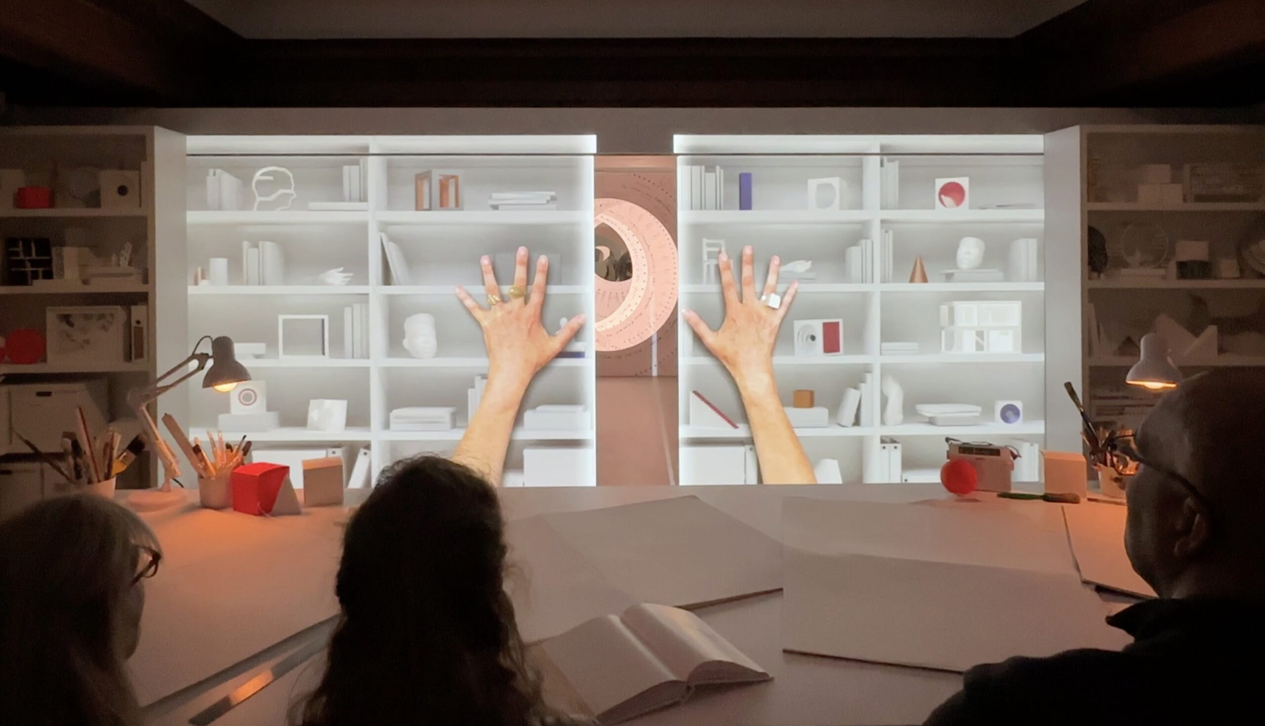 Three people sit and watch a wall split open down the middle. The wall is projected to look like the bookshelves in the room around it, with two larger-than-life hands pushing the wall open.