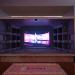 A small model of a theater inside a large wooden crate. The theater's stage is a screen showing a purple and pink Es Devlin stage design.