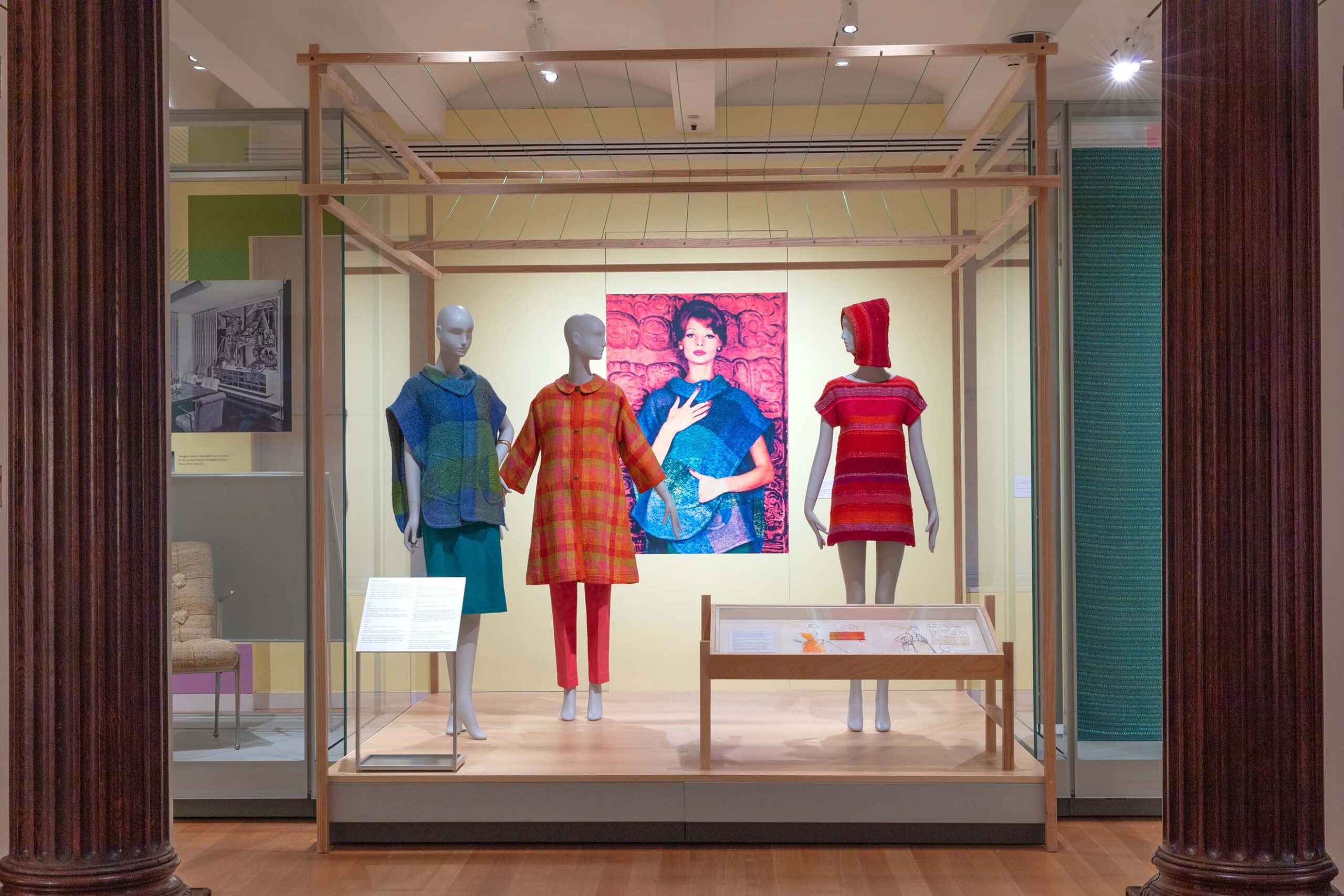 Three mannequins dressed in brightly covered textiles on view in front of a large photograph showing a woman dressed in similar textiles.