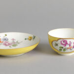 A delicate yellow and white cup and saucer set, decorated with intricate depictions of flowers.