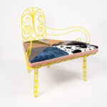 A chair with a yellow wire back and a disproportionately long cushioned seat upholstered with various patterned fabrics.