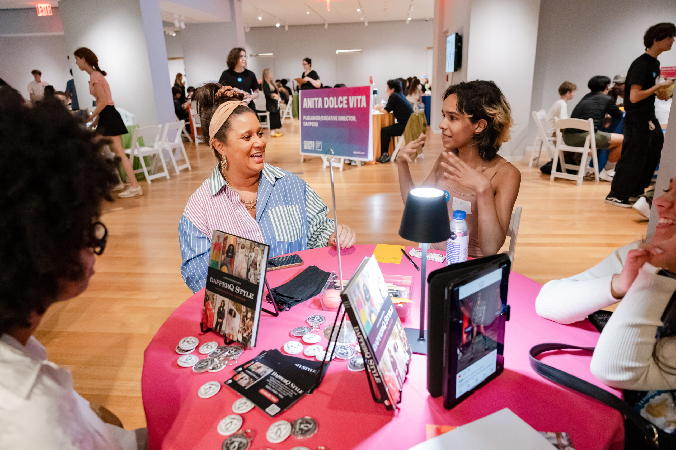 A photograph of a group of teenagers and an adult are sitting around a circular table and talking. The light- brown skinned adult who wears a colorful striped button down is smiling and in conversation with the light-brown skinned teenager with short brown hair sitting on their right. On the table is a small lamp, and books titled ‘ DapperQ Style’, and many key rings. Behind them are other circular tables that have small groups of adults sitting and talking.