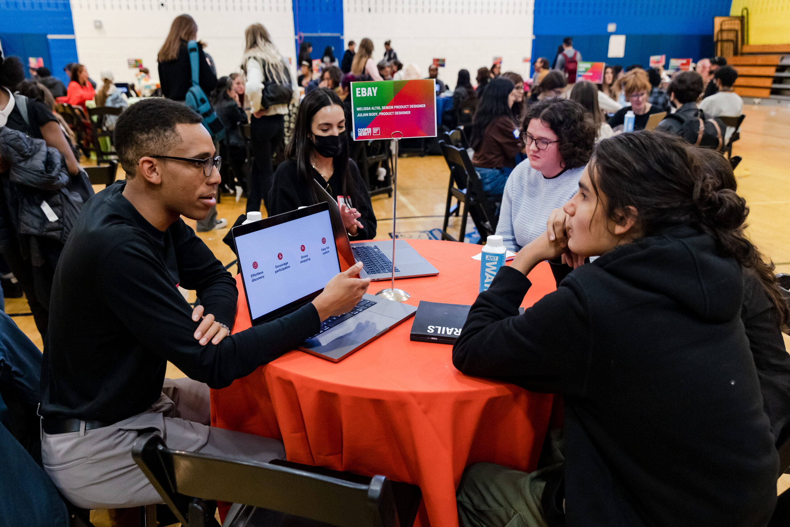 A photograph of a group of teenagers and an adult are sitting around a circular table and talking. The dark-skinned with closely shaved hair is talking to medium-skin toned teenager sitting beside them and referencing a slide on their laptop. Behind them are other circular tables that have small groups of adults sitting and talking.