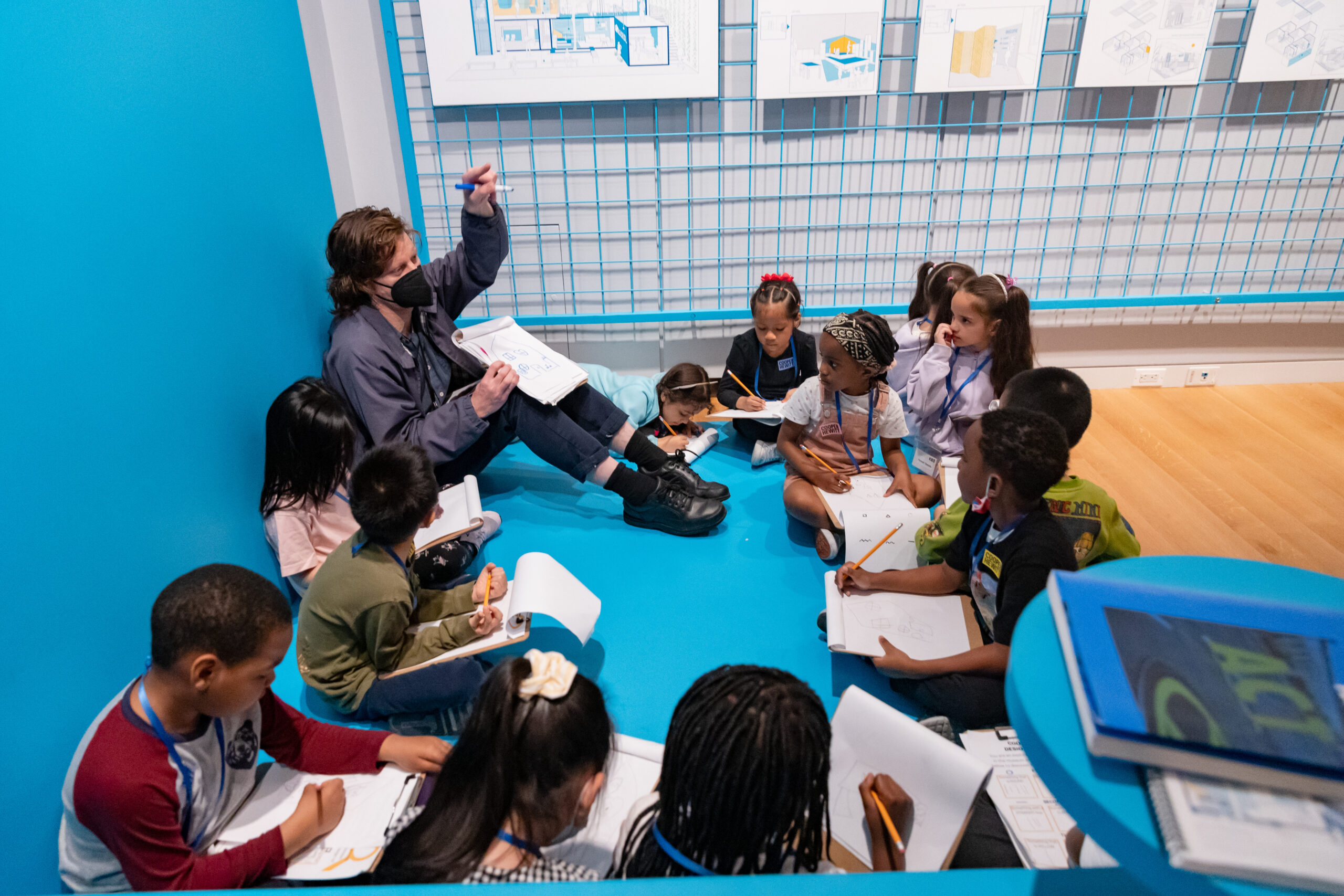 A photograph of an adult and group of children sitting in a circle on the ground in the corner of a room doing an activity. Each child has a clipboard and pencil and are listening to the adult. The light-skinned adult has short brown hair, is holding a clipboard with a drawing of a house and is pointing. The wall behind them is bright blue.