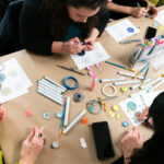 A photograph, taken from above, of a group of people sitting down at a table and painting colourful dots onto small circular objects the size of a quarter. Various art materials are scattered across the table, including markers, paintbrushes, stencils, and coloring pages with circular designs that mimic the real-life objects that people are painting.