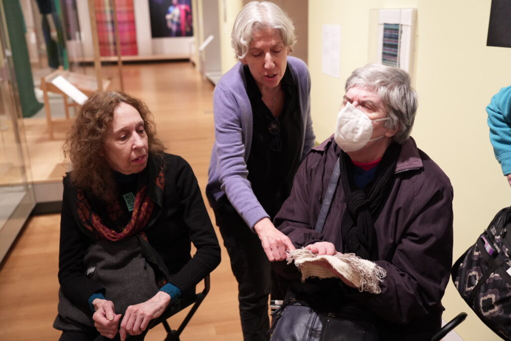 A photograph of two people sitting next to one another in a gallery space. The person to the right is holding a white and grey textile sample with loose threads. A person stands in the middle behind the two people sitting, their hand reaches across the person sitting on the right and touches the textile.