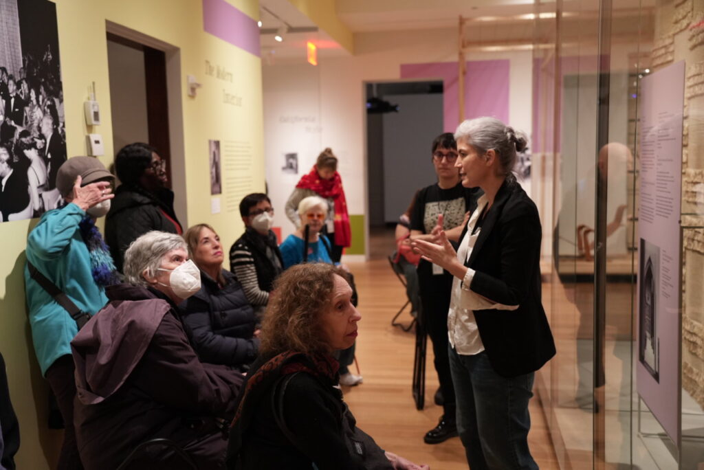 A photograph of a group of adults sitting in a gallery space. An adult stands in front of the group, speaking next to the objects on display. The person speaking has grey hair tied in a bun and is wearing a white button-down shirt and black sports jacket.