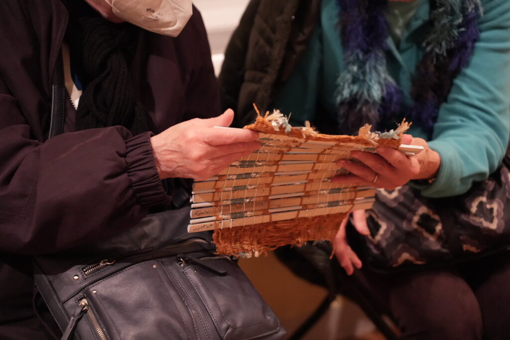 A close-up photograph of two people’s hands holding a textile sample. The textile sample has orange, grey, and silver strands going in a vertical direction. The weave of the yarn holds together multiple bamboo sticks that sit side by side going in a horizontal direction.