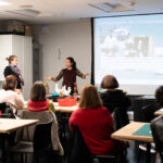 A photograph of two adults giving a presentation in front of a room of adults. The light-skinned adult with brown hair slicked back into a ponytail is speaking to the group. Behind them is a large screen titled, “Conservation: an introduction”, that has multiple images of people doing conservation work. In front of them is a table of various decorative objects that look like vases or candle holders.