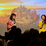 A photograph of two women holding microphones and stand in front of an audience giving a presentation. The light skinned woman with long straight black hair speaking on the right is Es Devlin, she wears a yellow sweater and a chunky necklace. The light-skinned woman with short brown hair on the left is looking and smiling at Es Devlin. Behind them is a screen projecting an image of a stage composed of large-scale hands and playing cards that appear to be thrown into the air. The stage is set on a waterfront, and in the background the sun is setting.
