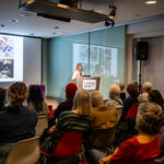 A photograph of a woman with shoulder length grey-blonde hair and a white knitted cardigan stands at a podium speaking in front of an audience. Behind her is a screen projecting a slide of multiple images, such as a color sample sheet, textile samples on a table, and a mood board.
