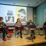 A photograph of four people sitting in chairs across a stage. The light skinned man sitting in the middle, Seymour Chwast, is speaking into a microphone, he has a grey beard, wears glasses, and a baseball cap. Behind the group is a large screen projecting multiple images of posters and illustrations that relate to War, each designed by Seymour Chawst.