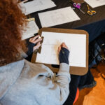 A photograph of a bird's eye view of a light skinned person with curly red hair sitting at a table and drawing an interior space on a piece of paper attached to a clip board.
