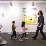 A photograph of a group of children walking in a single file line behind an adult in a gallery space. The adult is standing at the front of the line and is facing the children. The adult and children are all carrying a clipboard and pencil. On the wall behind them there are a series of graphic posters that show the winner of the national high school design competition.